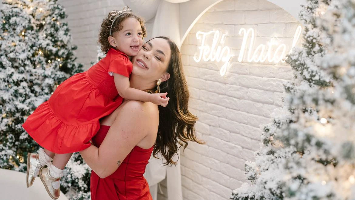 Mãe abraçando uma menina de vestido vermelho no cenário de Natal