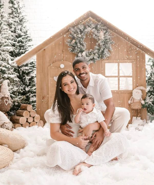 Família com um bebê, reunida diante de uma casinha de madeira decorada para o Natal