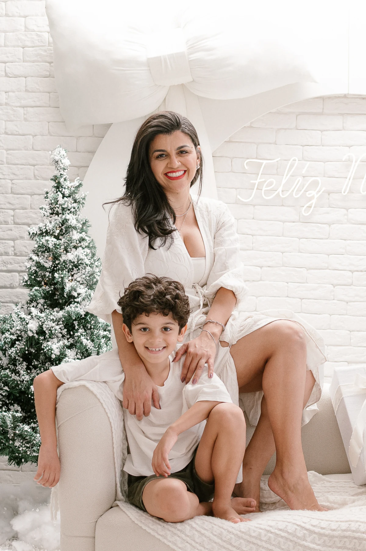 Thuanni Guimarães com uma criança em um cenário natalino branco, ao lado de uma árvore e um grande laço